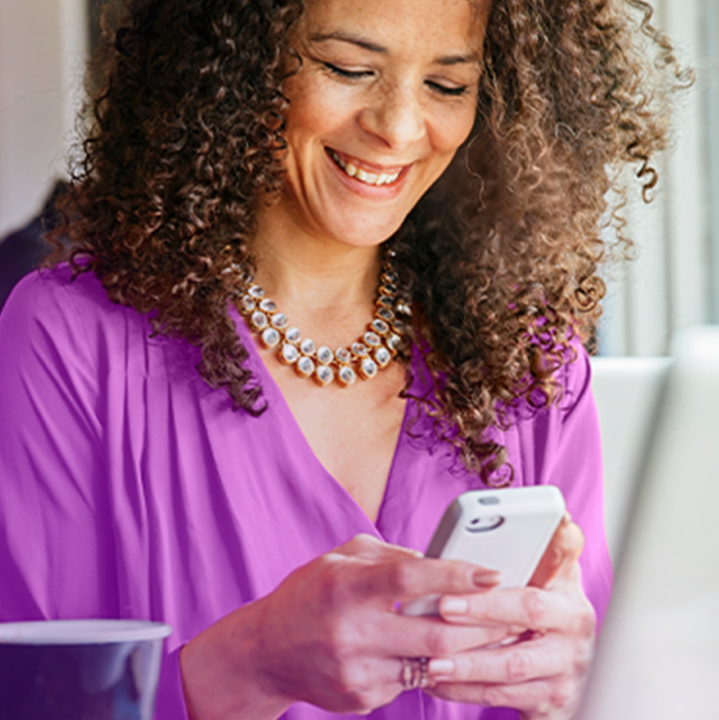 A woman with curly hair smiles as she looks down at her smartphone, holding it in both hands. She is wearing a vibrant purple blouse and a statement necklace.