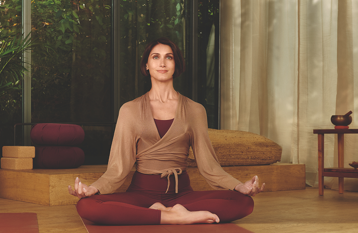 Woman sitting cross-legged on a yoga mat in a calm, softly lit room, meditating with her hands resting on her knees.