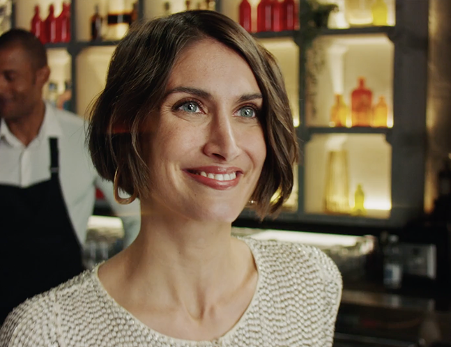 Smiling woman with short brown hair, sitting in a warmly lit room with decorative glass bottles in the background.